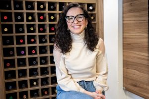 Sommelière Jessica Marinzeck sorrindo, sentada em frente a uma adega de vinhos. 