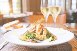 Prato branco com salada de camarão, e taças de vinho branco ao fundo.